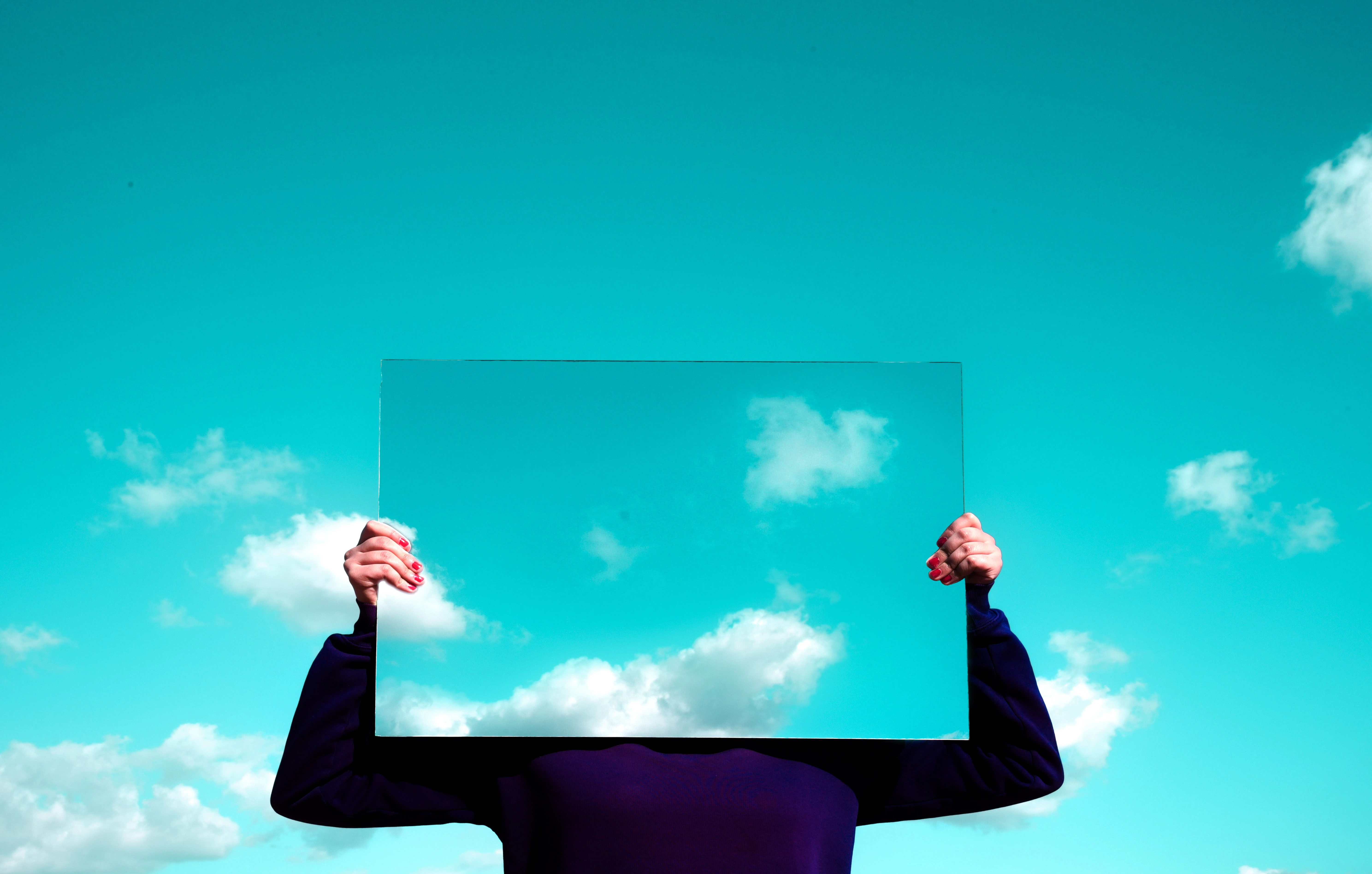 blue sky with clouds behind man in black jumper holding up mirror reflecting the sky and clouds