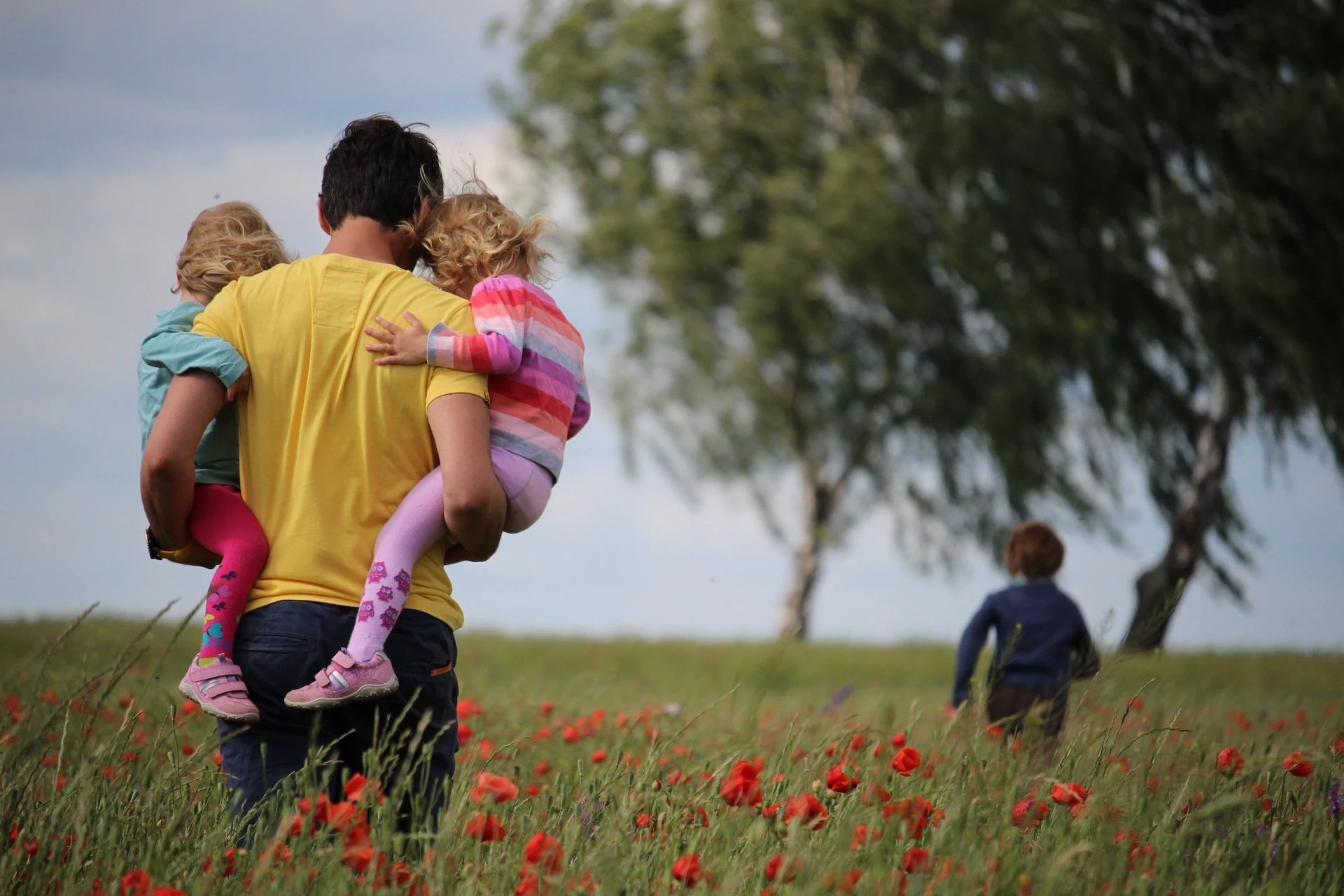 man in yellow shirt holding two children, walking through field of poppies