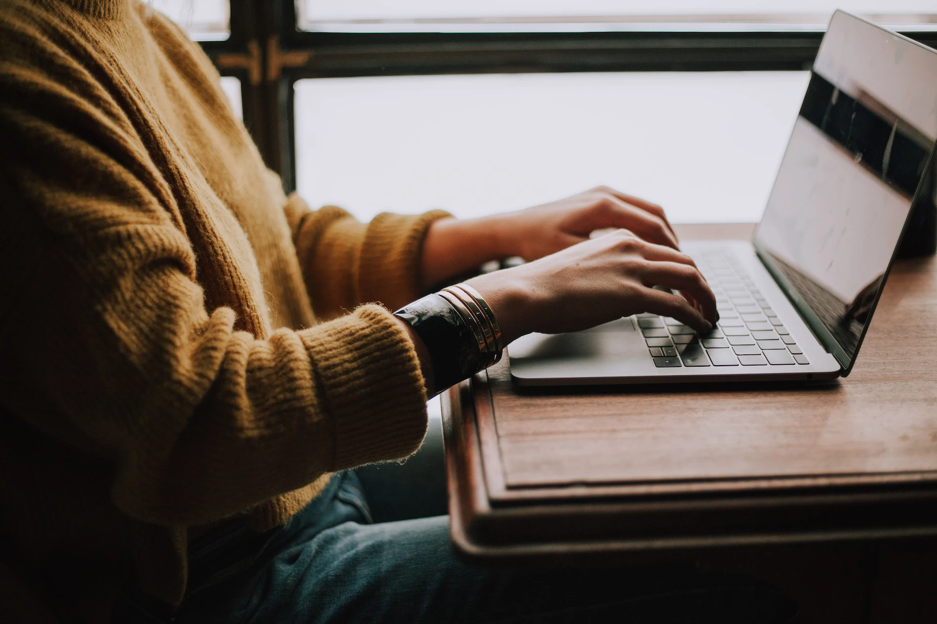 person wearing brown jumper typing on laptop sitting at wood desk