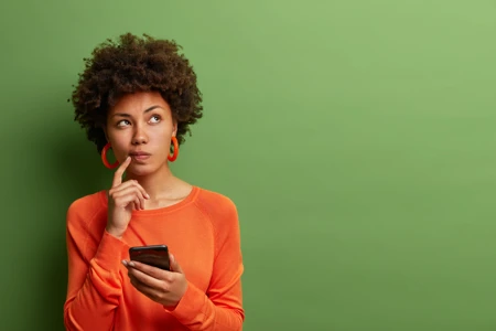 Photo of pretty ethnic woman ponders on how to answer question, thinks deeply about something, uses modern mobile phone