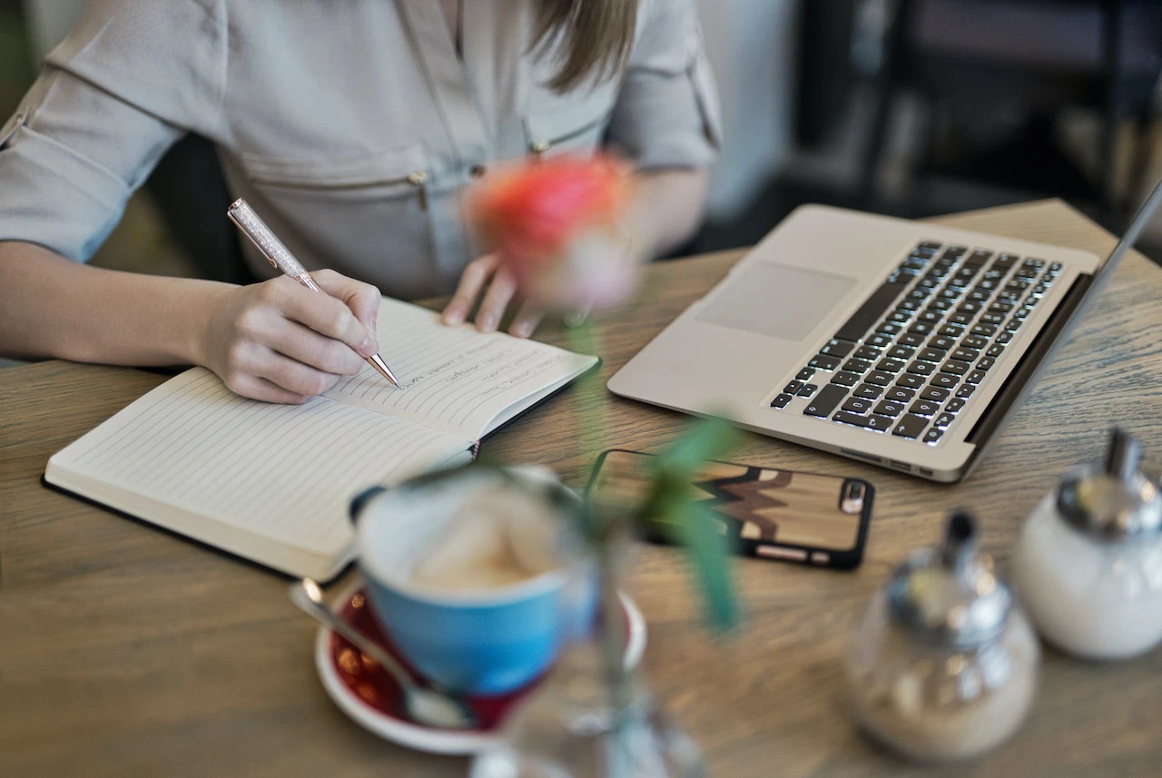 person writing in a notebook with a laptop and hot drink in front of them