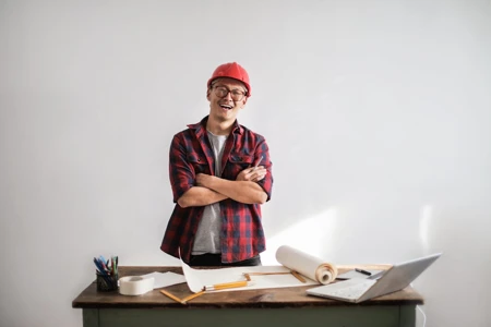 A smiling construction worker in a hardhat and glasses, standing in front of a blueprint.