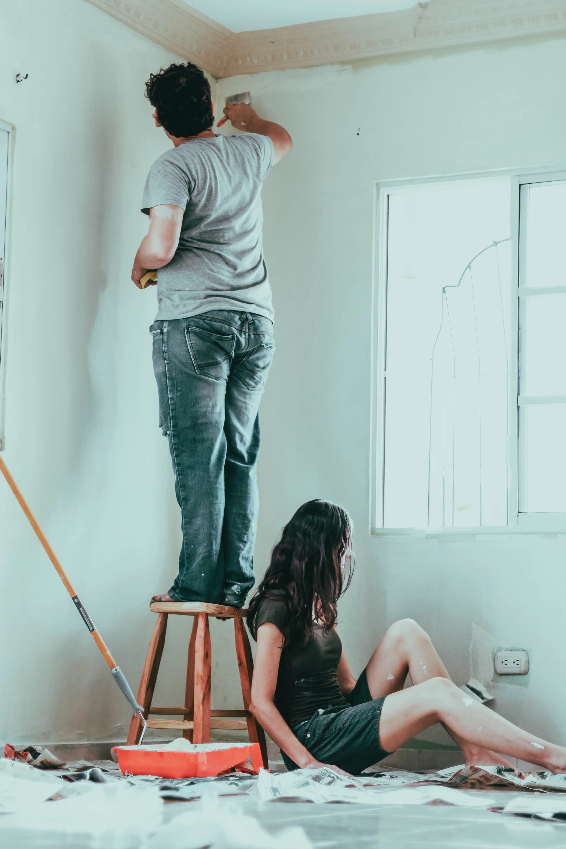 two people painting a room white - man standing on stool to the left, woman sitting on the floor