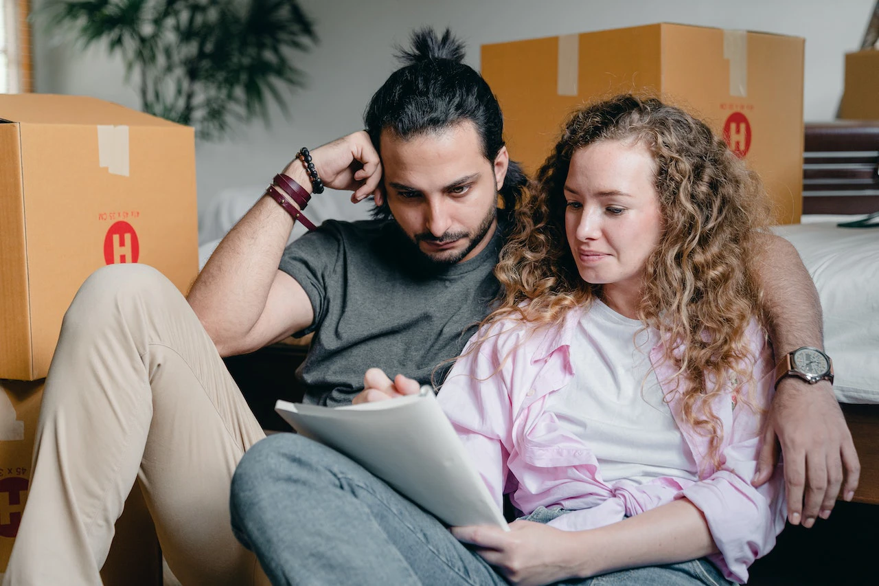 couple looking at paper surrounded by cardboard boxes - home improve or move