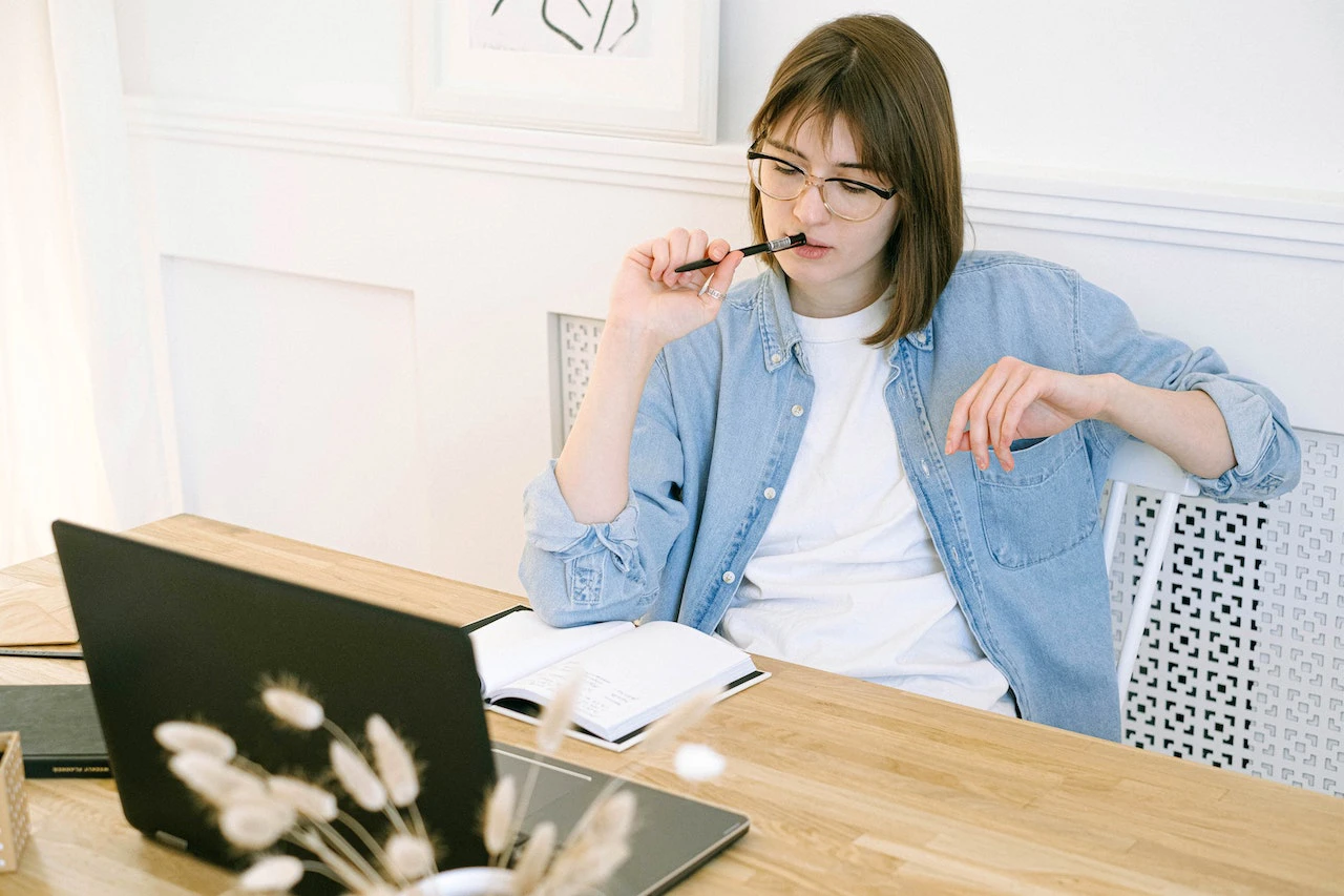 Woman looking at laptop and notebook while holding a pen to her mouth