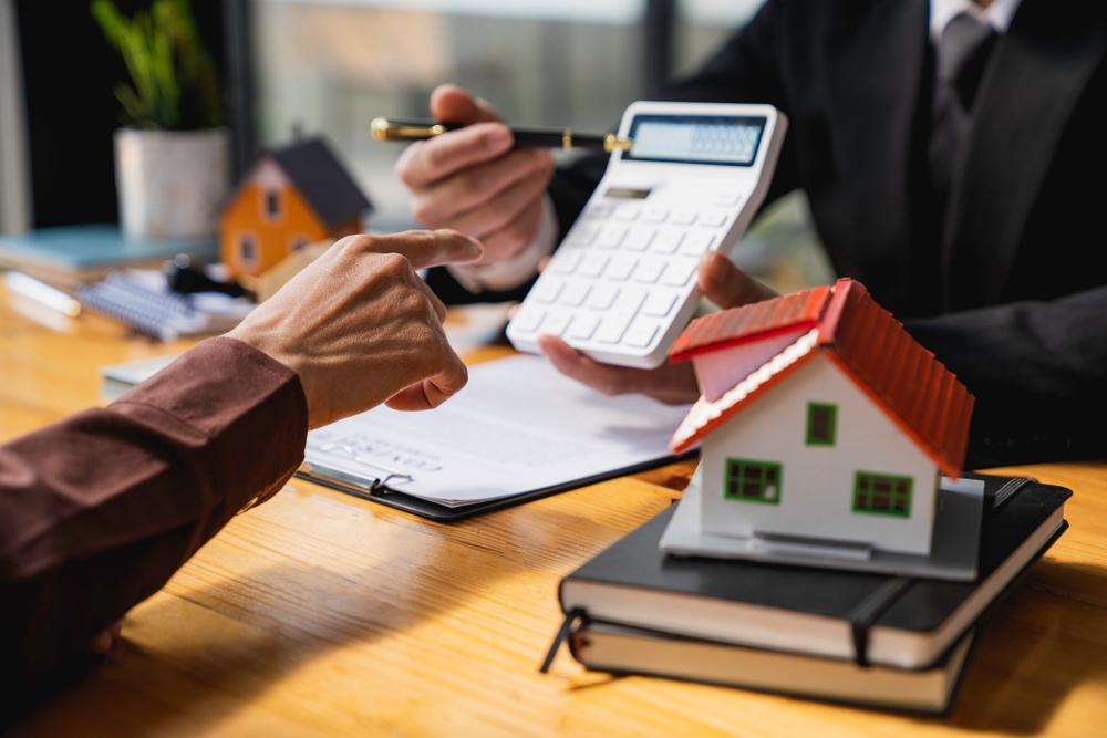 person sat on one side of a desk showing another person a figure on a calculator, with a model house on the desk