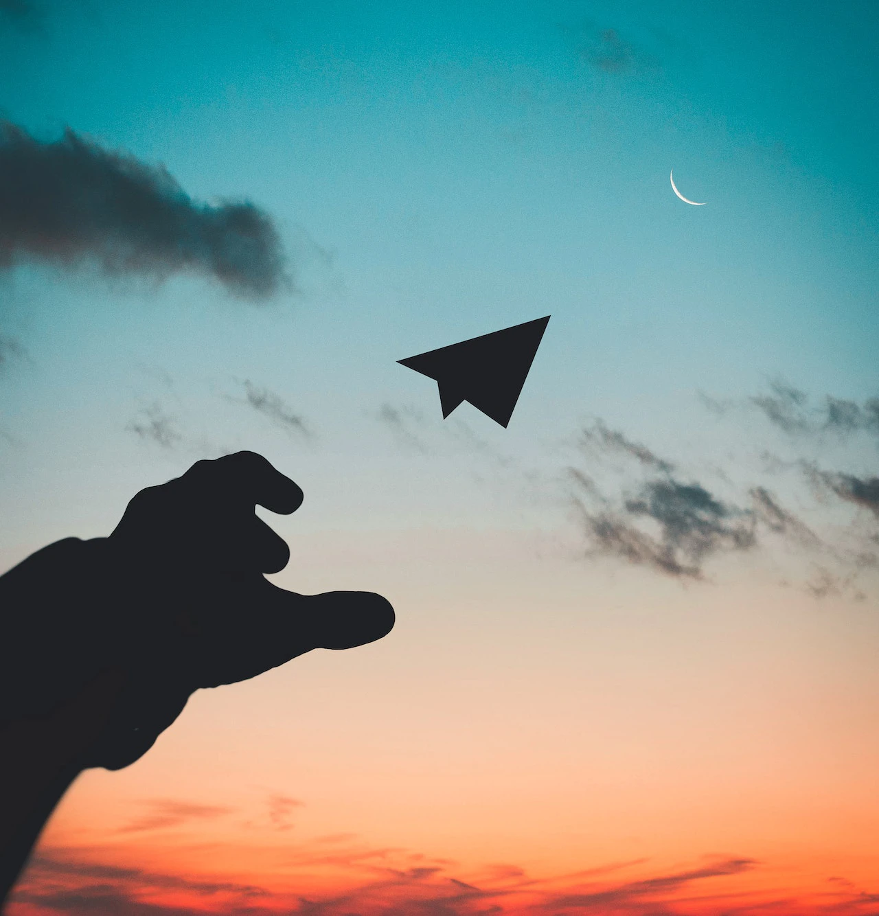 silhouetted hand throwing a paper airplane towards a crescent moon against a sunset sky