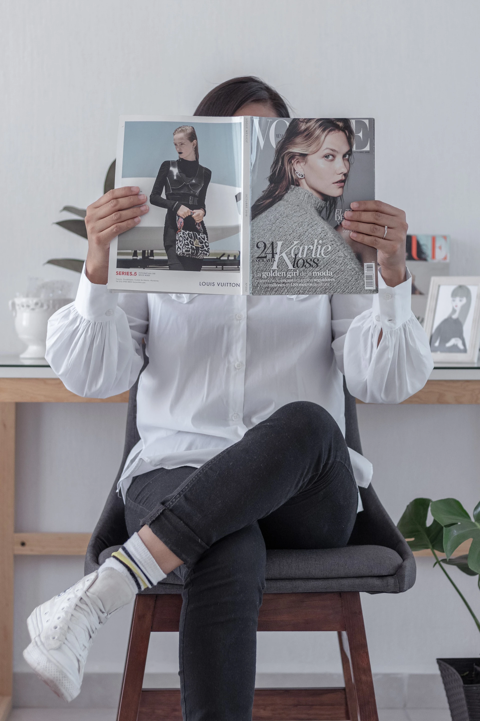 woman sitting down wearing a white shirt, jeans, and trainers, reading a fashion magazine