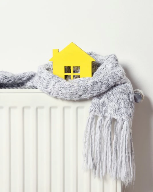 House model wrapped in scarf on radiator indoors. Winter heating efficiency