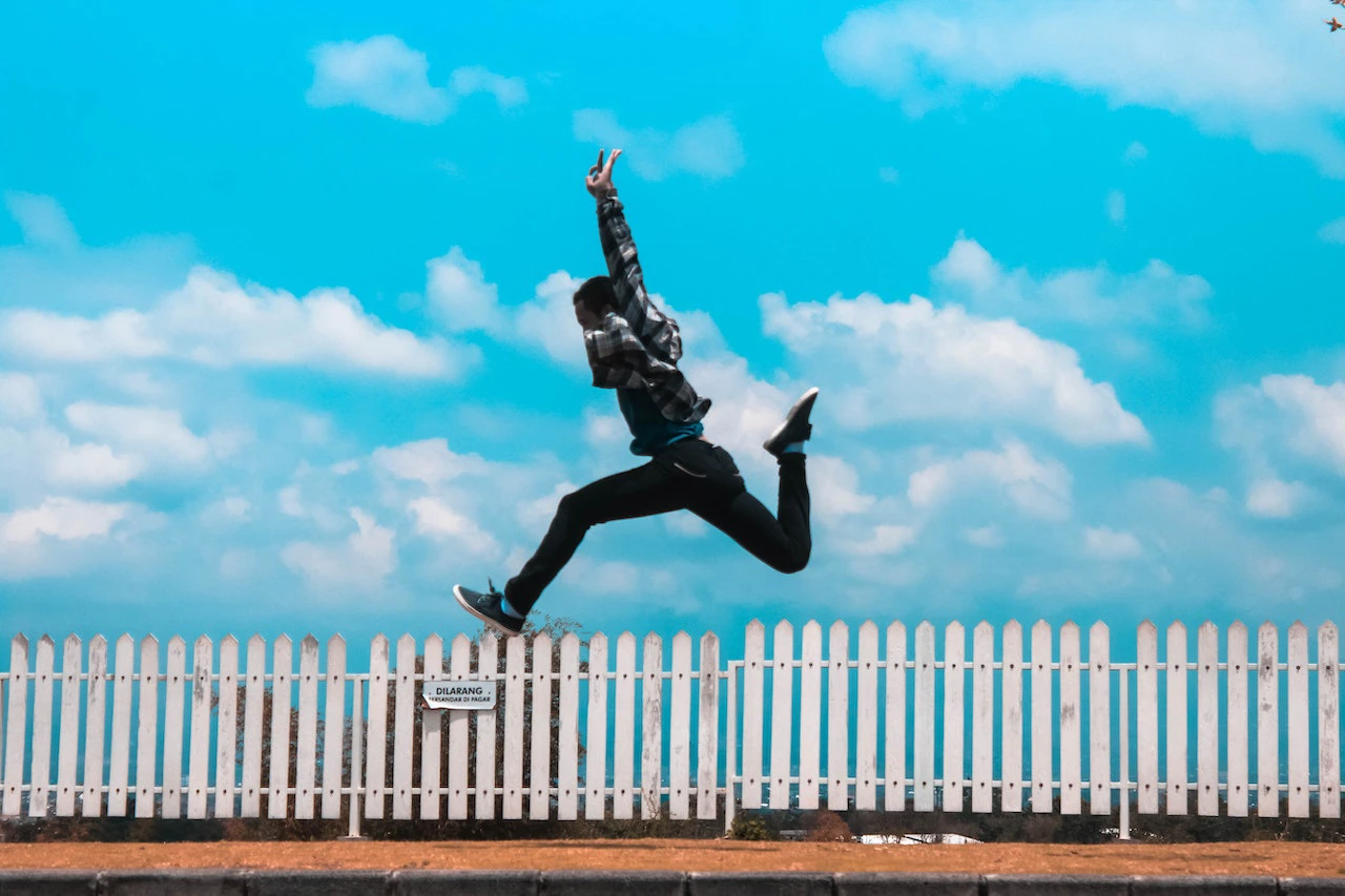 man in dark clothes jumping above a white picket fence against a bright blue sky with puffy white clouds