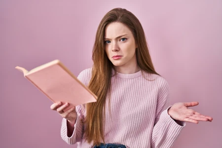 Young caucasian woman reading a book over pink background clueless and confused expression