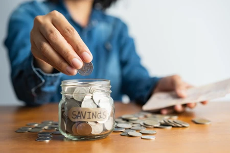woman hand is putting a coin in a glass bottle and a pile of coins on a brown wooden table